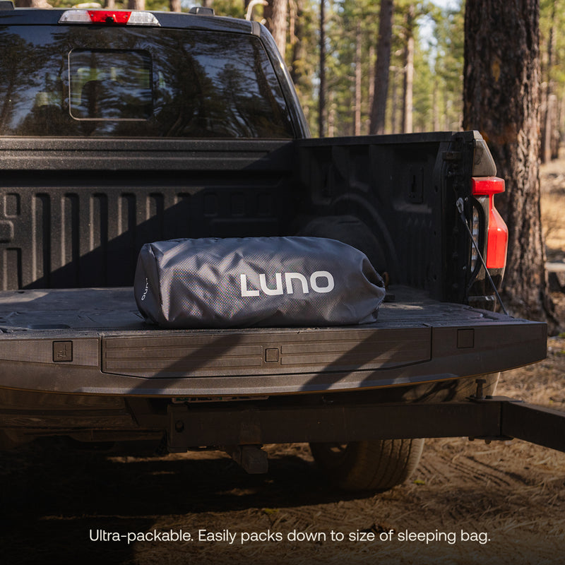 A Luno® AIR Truck Bed Mattress sits on the open tailgate of a black pickup in a forest, showcasing ultra-packable design that easily compresses for comfortable truck bed sleeping wherever you go.