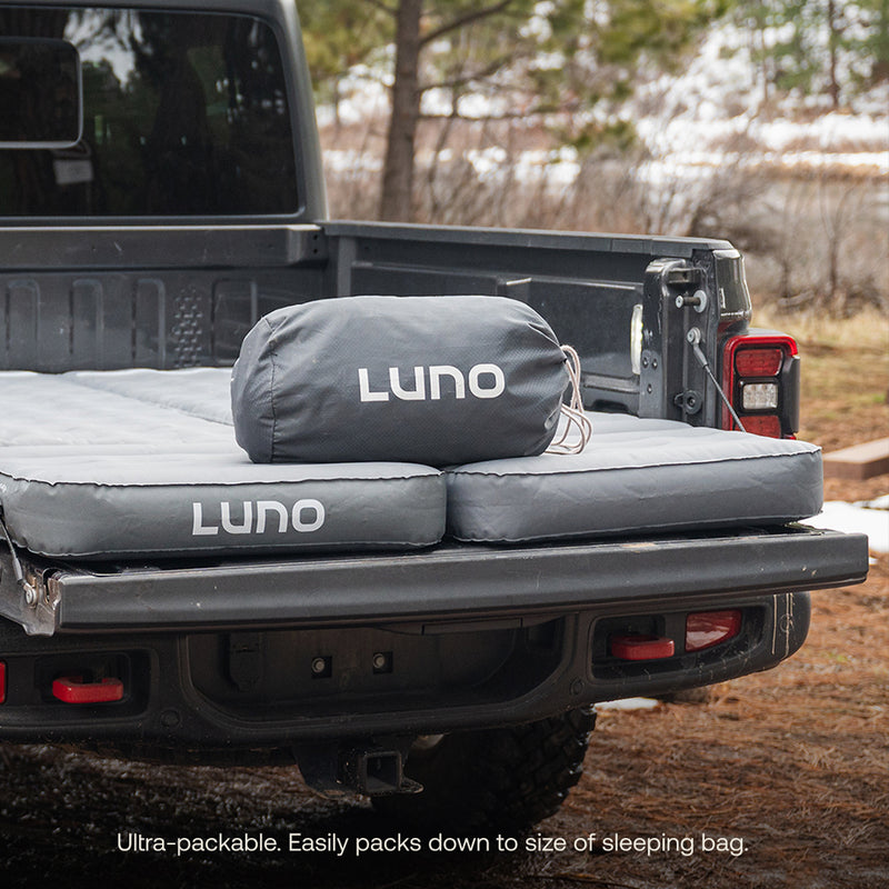 Two gray Luno® AIR Truck Bed Mattresses and a packed Luno bag are set up in a pickup truck bed outdoors among trees. Text: “Ultra-packable. Easily packs down to the size of a sleeping bag.”.