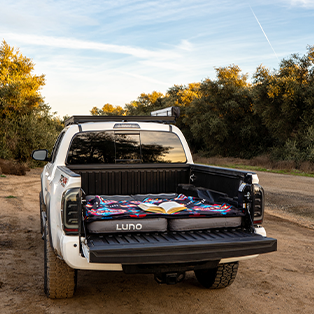 A white pickup truck with its tailgate down is parked on a dirt road surrounded by trees. The truck bed features an inflated Luno air mattress with a colorful blanket and an open book placed on top. The scene suggests a camping or outdoor adventure setting.