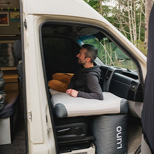 A person sits comfortably inside a camper van, resting on an inflated mattress with "Luno" written on it. The camper van interior is visible, with a cozy setting that includes cabinets and various seating areas. The person appears relaxed, surrounded by nature.