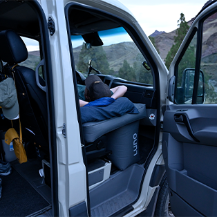 A person reclines in the driver's seat of a white vehicle, facing away from the camera. The vehicle door is open, revealing the interior. There are trees and mountains in the distant background.