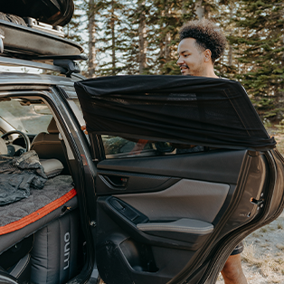 A person adjusts a sunshade on the rear door of a parked car in a wooded area. Camping gear, including a mattress and a sleeping bag, is visible inside the vehicle. The person is smiling and appears to be preparing for an outdoor activity.