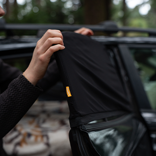 A person is installing a black car window shade on an open car door. The window shade has a small yellow tag. The background shows a blurred wooded area, indicating an outdoor setting.