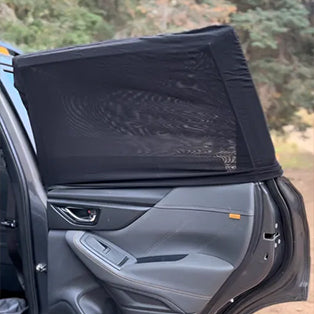 A close-up of the inside of a car door with a black mesh sunshade covering the window. The door is open, revealing the interior, including the armrest and control panel. Trees and greenery are visible in the background.