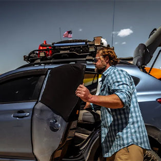 A man in a checkered shirt is opening the back door of a gray SUV. The vehicle is equipped with various gear on the roof, including an American flag. The background is a clear sky with a few clouds.