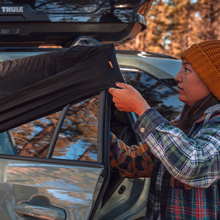 A person in a plaid jacket and orange beanie is setting up a cover on the window of a parked car with a roof storage box, surrounded by trees.