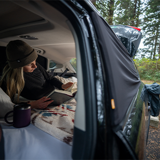 A person wearing a beanie and sweater lies in the back of an SUV, reading a book. The car's hatch is open, showing a cozy setup with blankets and a mug, surrounded by a forest scene.