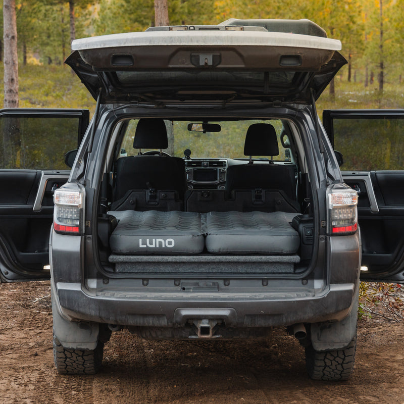 A Luno® AIR 4Runner Vehicle Mattress is set up in the cargo area of an SUV, which is parked in a forest with its rear hatch and both rear side doors open. The sturdy, durable fabrics of the cargo area complement the comfort provided by the inflated mattress, making it ideal for car camping. Trees and foliage are visible in the background, ensuring a perfect blend of comfort and nature.