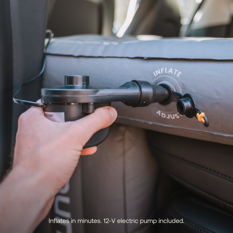A person's hand holds the Luno® AIR Vehicle Mattress's 12-V electric pump, inflating the mattress in a car. The pump nozzle is inserted into the mattress's "Inflate" valve. Text on the bottom reads, "Quick setup. Inflates in minutes." Durable fabric and 12-V electric pump included.
