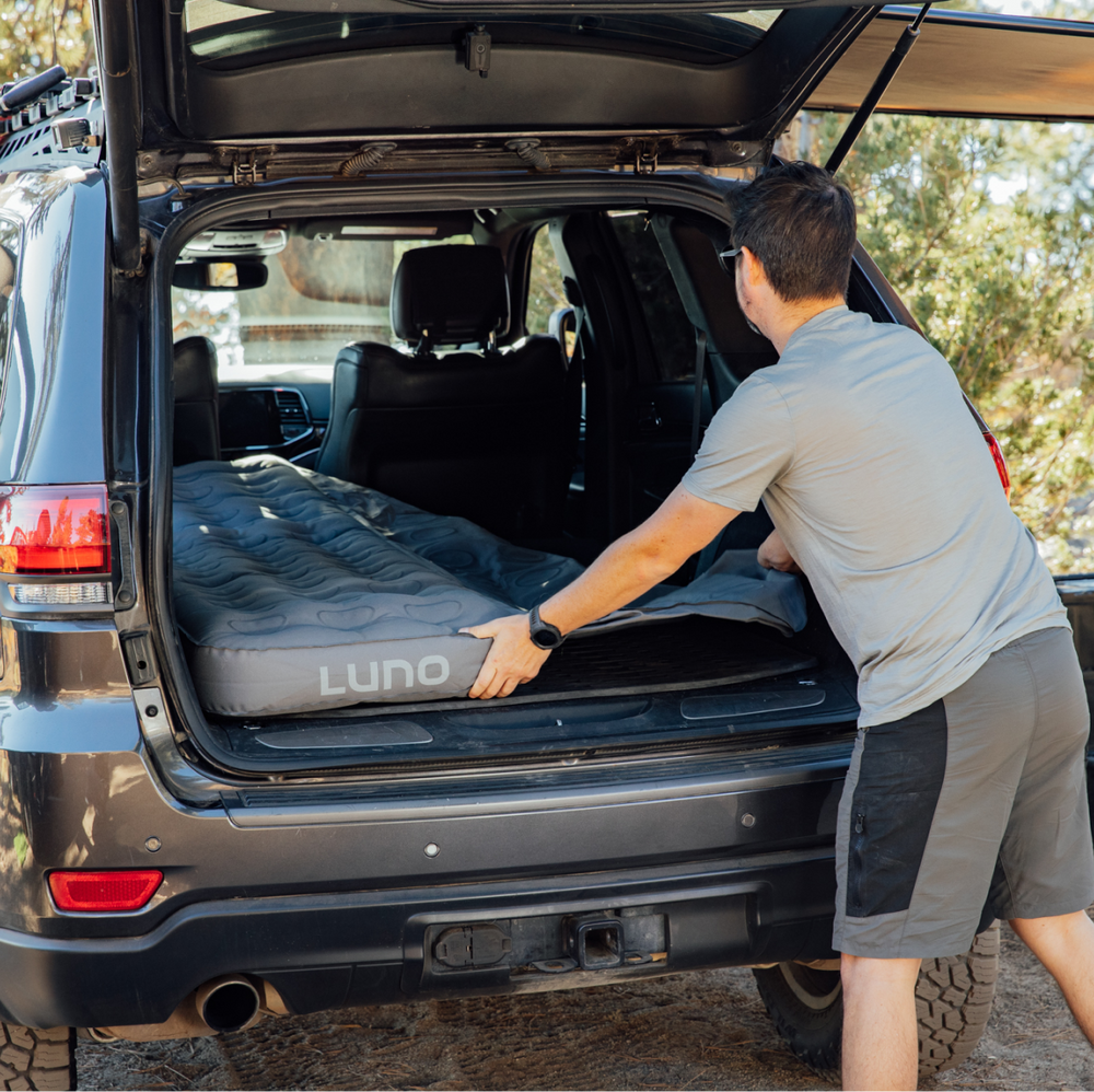 A person is setting up an inflatable mattress labeled "Luno" in the back of an SUV. The SUV's rear hatch is open, revealing the spacious interior. The scene suggests preparations for camping or sleeping in the vehicle, situated outdoors with trees in the background.