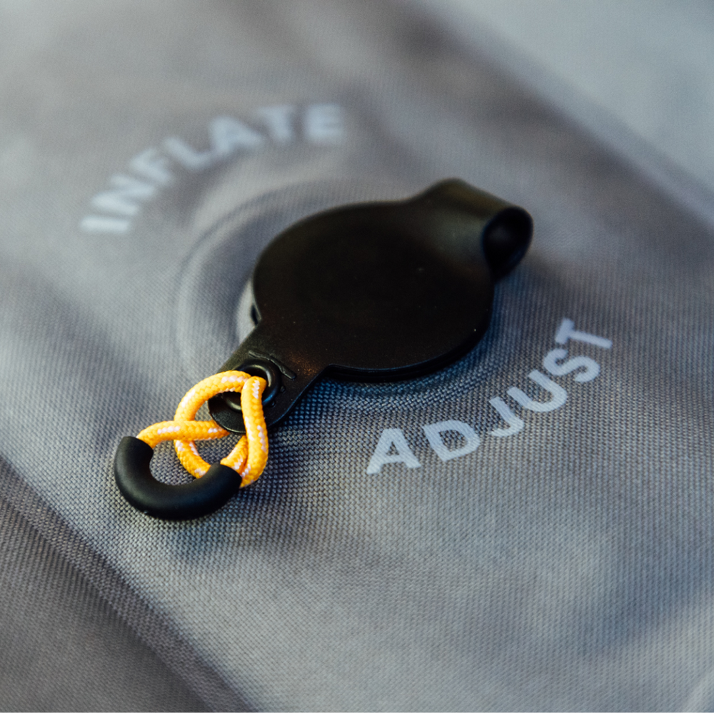 Close-up of a black circular valve with a small black handle on a grey fabric surface. The words "INFLATE" and "ADJUST" are printed around the valve, and a yellow cord is looped through a hole in the handle.