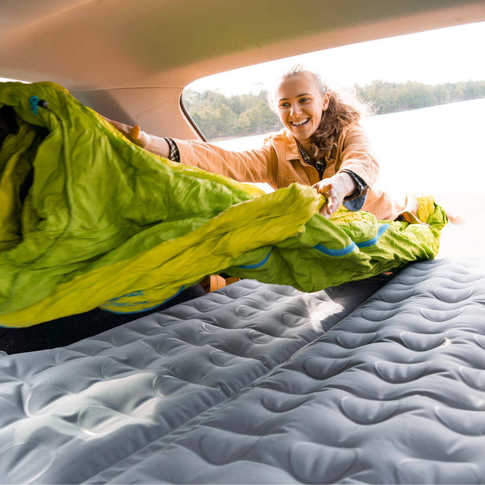 A person with a ponytail, wearing a beige jacket, is smiling and laying out a green sleeping bag inside a car with a blown-up air mattress in the back. Trees and a lake are visible through the car's open back hatch.