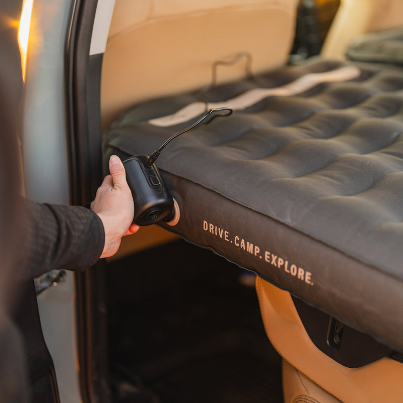 A hand uses an Inflatable Air Pump to inflate a mattress inside a vehicle labeled with the text DRIVE.CAMP.EXPLORE.