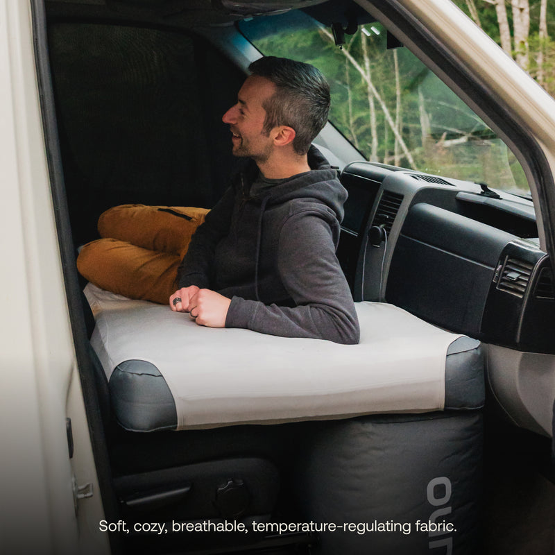 A man sits comfortably inside a vehicle, smiling and leaning against the backrest. He is resting on a Luno® AIR Van Front Cab Mattress covered with a soft, breathable, fitted sheet. A cozy, forested landscape is visible outside through the open car door. Text reads: "Soft, cozy, breathable, temperature-regulating fabric.