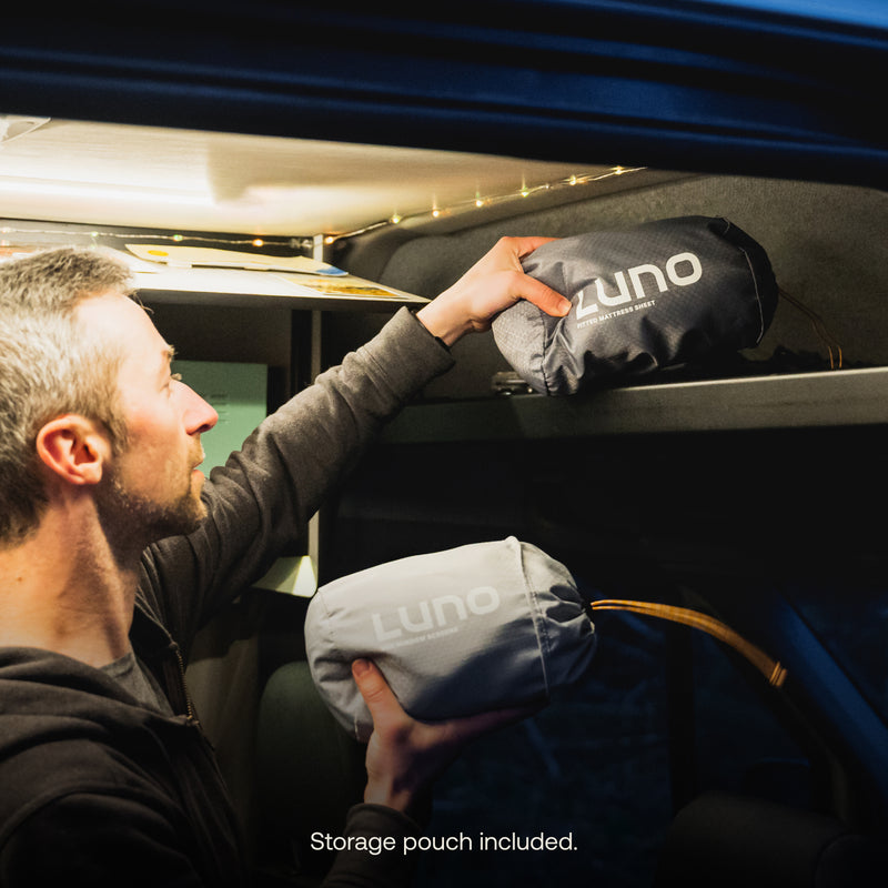 A person is placing two rolled-up Luno® AIR Van Front Cab Mattress Fitted Sheets, one black and one gray, onto a shelf in a dimly lit space inside a vehicle. The caption reads "Storage pouch included," ensuring you're ready to easily store your Luno® AIR Van Front Cab Mattress Fitted Sheet anywhere.
