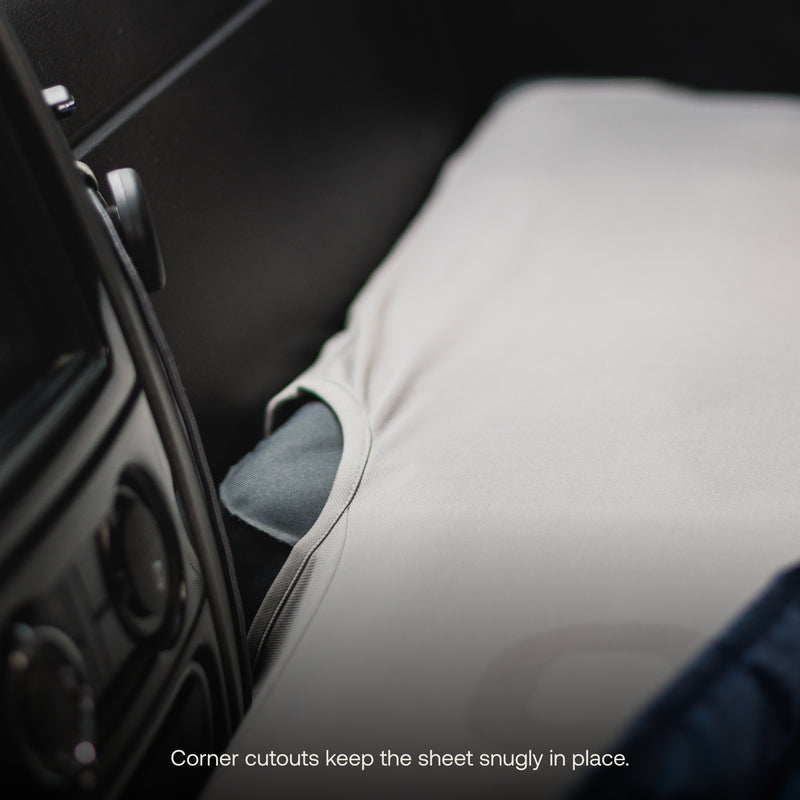 A close-up of a car seat covered with the AIR Van Front Cab Mattress Fitted Sheet from Luno®, featuring corner cutouts that ensure a snug fit. The Luno Air Mattress is partially visible on the left side, alongside the car's dashboard and air vent. Text at the bottom reads, "Corner cutouts keep the sheet snugly in place.
