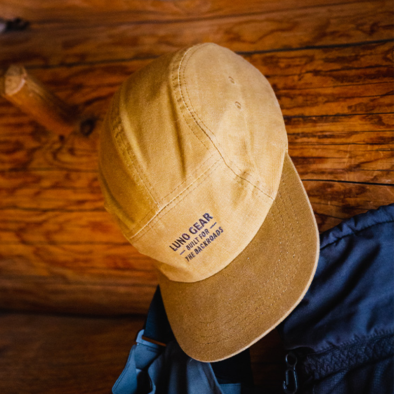 A yellow Luno HQ Backroads Canvas Hat with "Living Gear Built for the Backroads" printed on the front hangs on a wooden wall. Crafted from waxed cotton canvas, its five-panel styling features a brim in a slightly darker shade of yellow. Below the cap, part of a dark-colored jacket is visible.