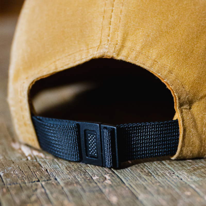 Close-up of the back of a mustard yellow Luno HQ Backroads Canvas Hat, showcasing its black adjustable strap and buckle on a wooden surface. This hat features waxed cotton canvas in five-panel styling for durability and style.