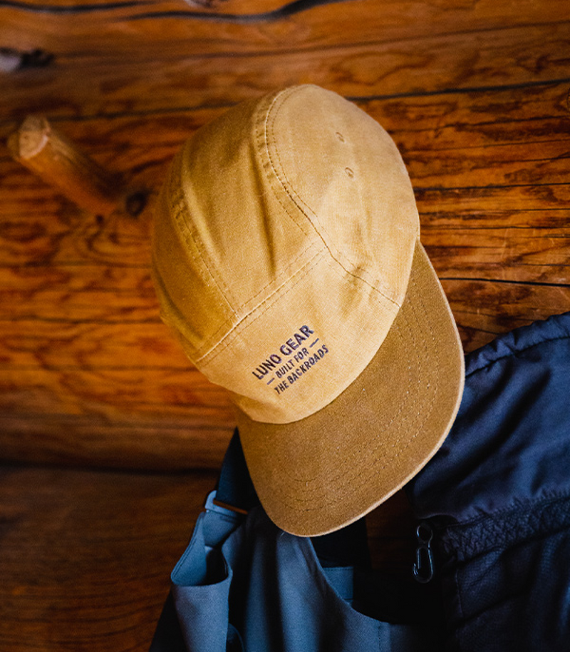 A beige and brown baseball cap hangs on a wooden wall. The cap has text that reads "WAND GEAR ANTOR AD THE BACKROADS" on the front. The background features a rustic, varnished wood surface and part of a black garment is visible below the cap.