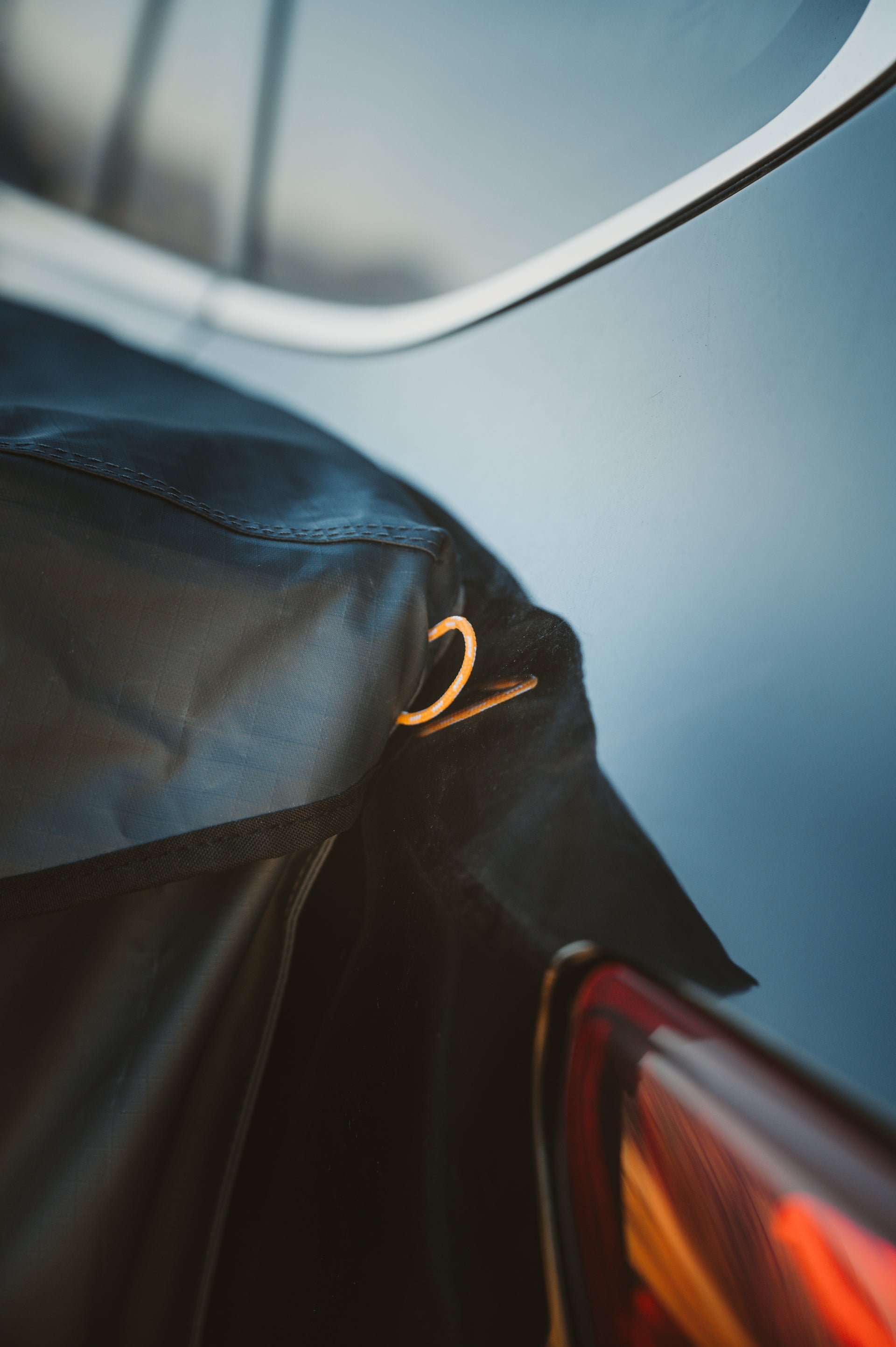 Close-up of a car's rear section showing part of a black protective cover attached to it. The cover is secured with a metal ring, and the red taillight of the silver vehicle is partially visible. The background is slightly blurred.