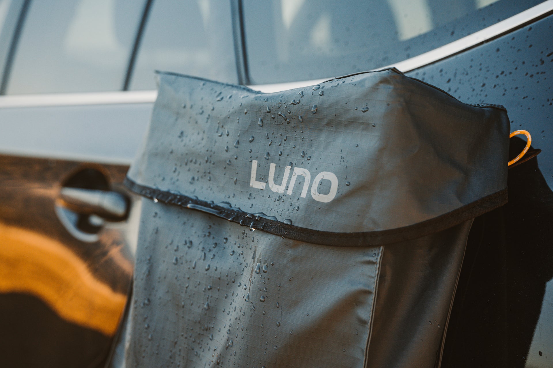 A close-up of a black car with a black waterproof bag labeled "LUNO" hanging on the window. The bag is covered in water droplets indicating recent exposure to rain or moisture. The car window is partially visible in the background.