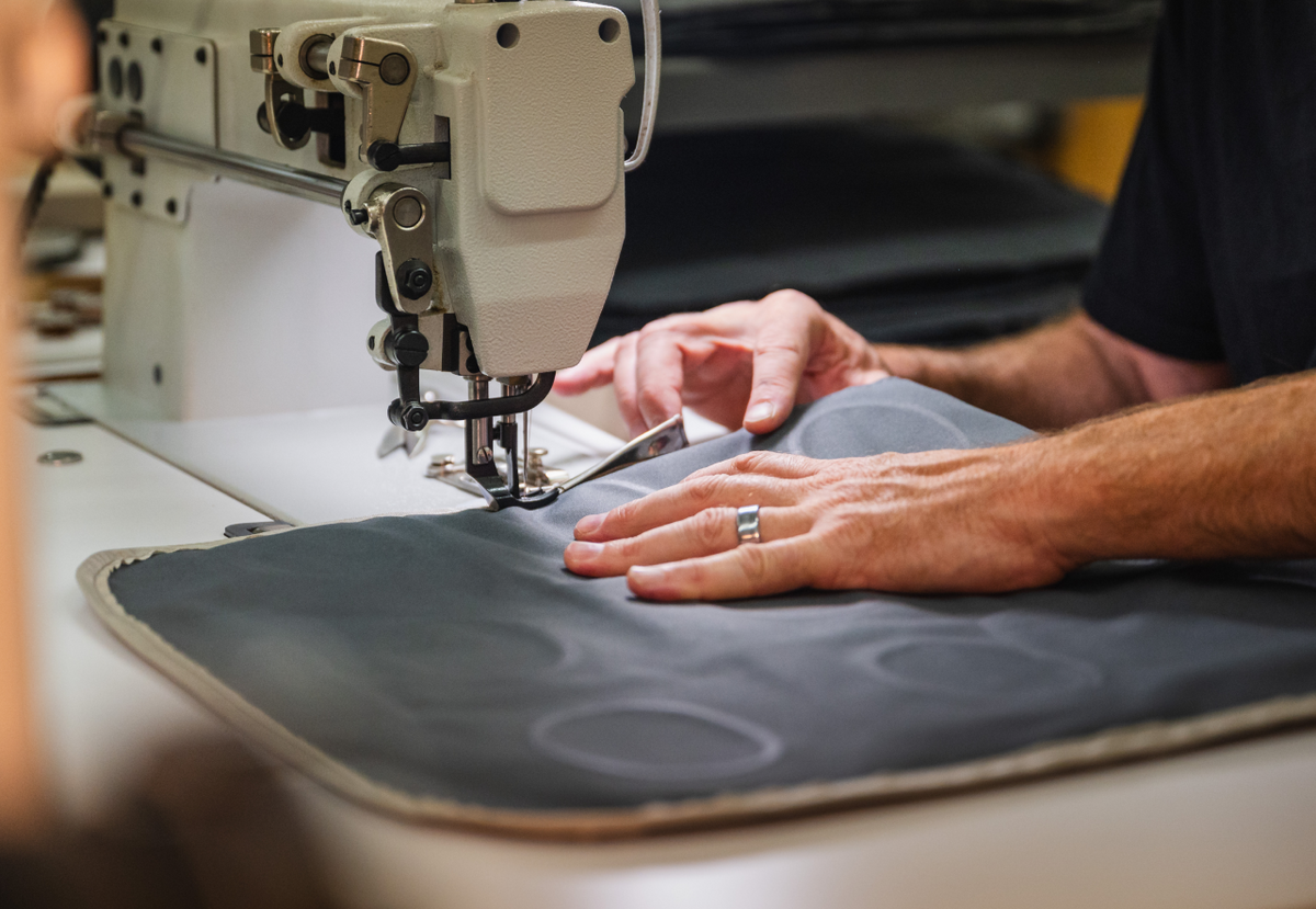 Close-up of a person's hands guiding a piece of dark fabric through a sewing machine. The machine has various knobs and gears visible, and the person is wearing a ring on their finger. The fabric appears to be thick and structured.