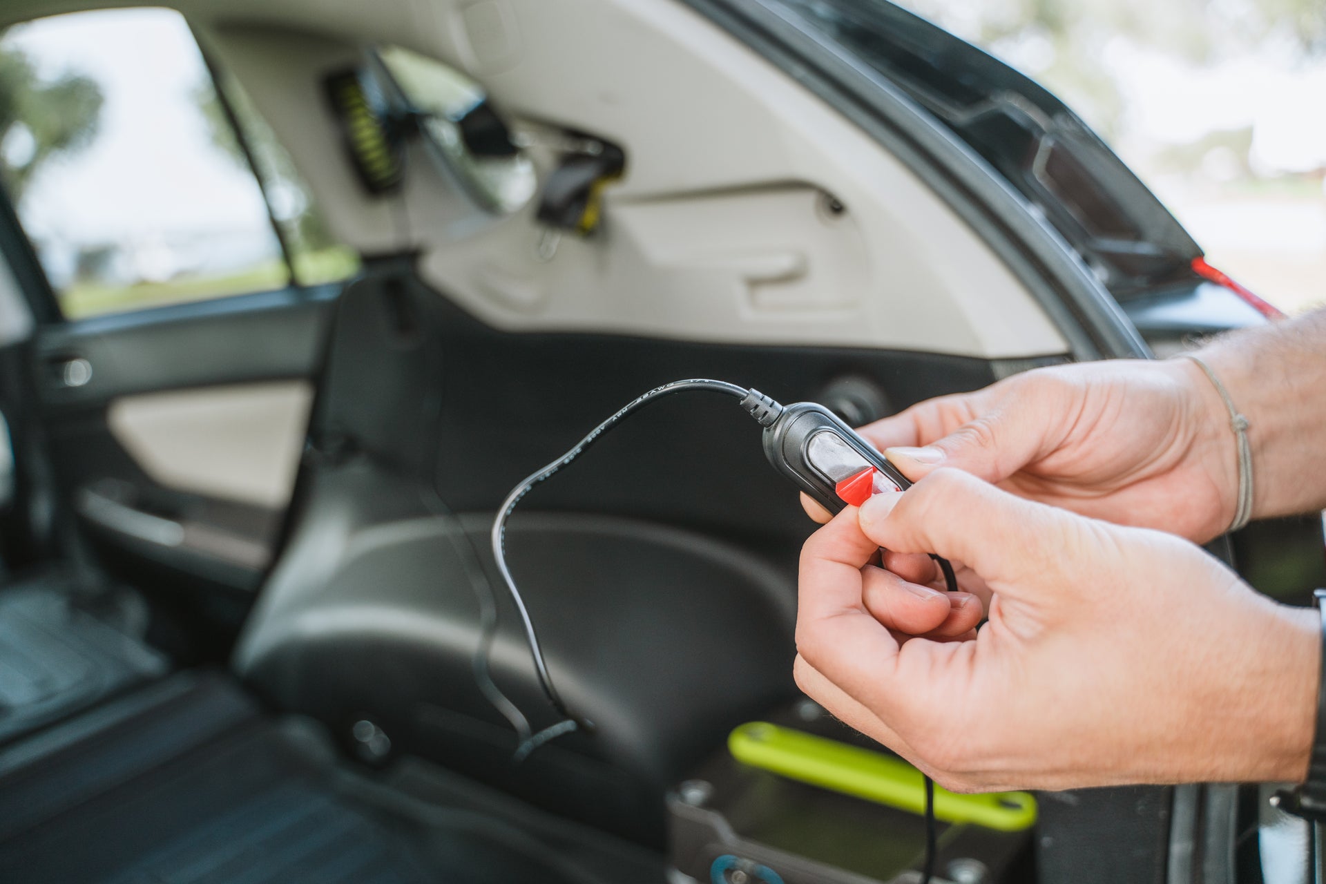 A pair of hands holds a black car charger plug with a red button inside a vehicle. The car's back door is open, revealing the interior including the back seat and trunk area. The scene appears to be outdoors with some greenery visible through the car windows.