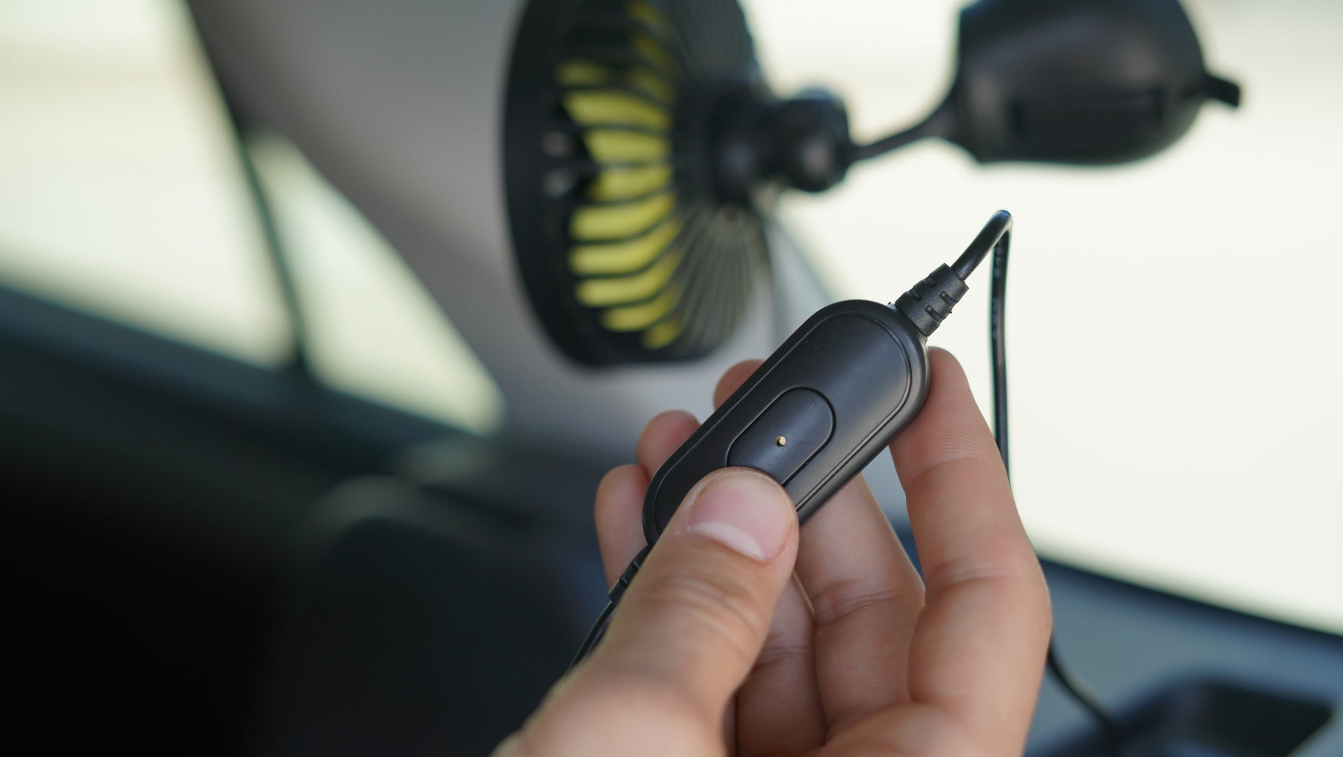 A close-up image of a hand holding a small black remote switch connected to a cable that leads to a black and yellow fan attached to the interior of a car. The scene appears to be set inside a vehicle.