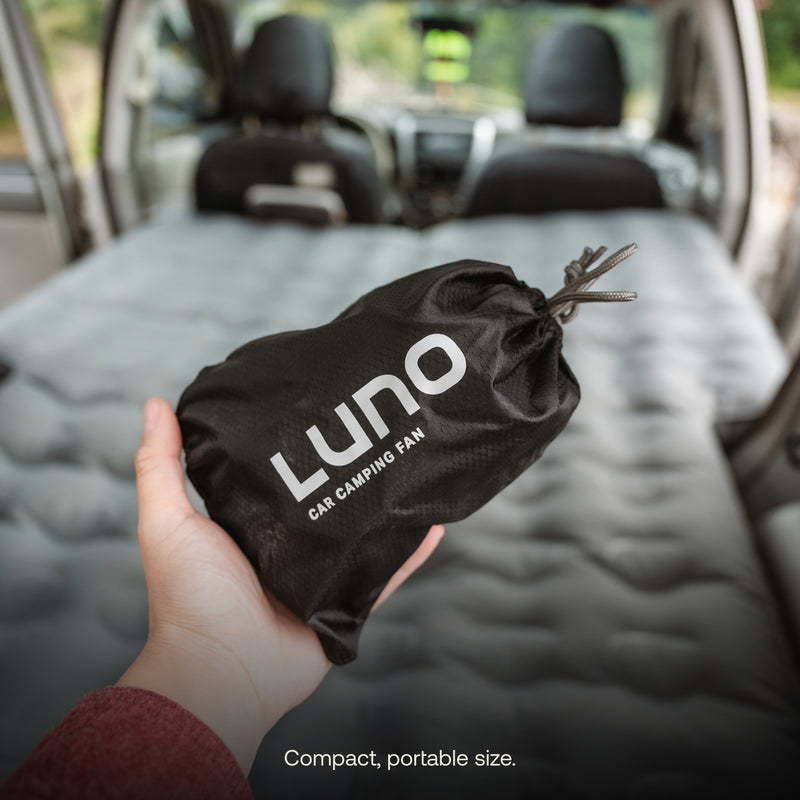 A hand holds a black drawstring bag labeled "Luno® Car Camping Fan" inside a vehicle equipped with an air mattress that covers the back seats. The setup hints at a compact USB-powered fan tailored for car camping. The text at the bottom reads, "Compact, portable size.