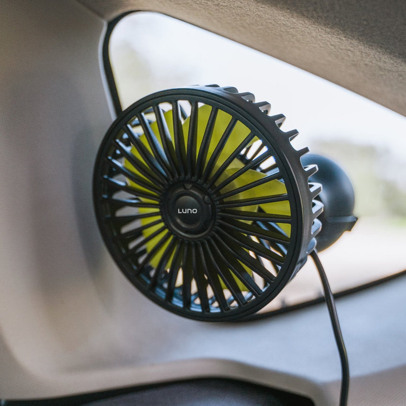 A pre-owned Camping Fan by Luno HQ, featuring a circular design, is mounted on a car window, offering a view of the outside. The fan's blades are enclosed in a protective grill and it is connected with a visible cord, all while maintaining like-new condition.