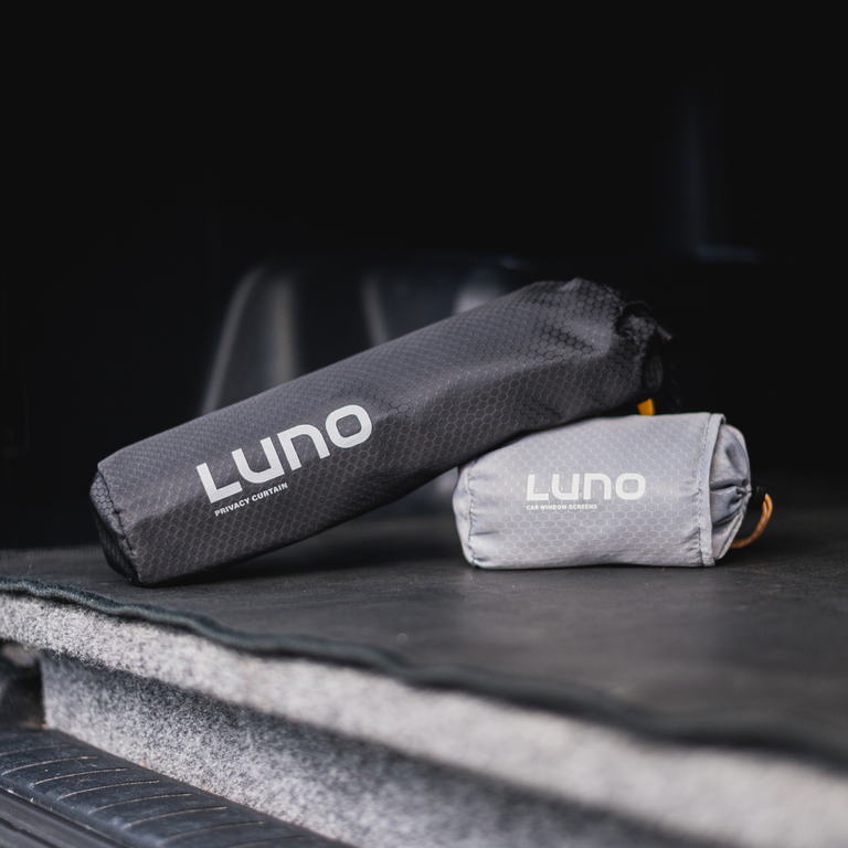 Two rolled-up Car Camping Privacy Bundles by Luno® are placed on a car's cargo area. The bundles are neatly packed, with one in a dark, textured bag and the other in a light gray bag, each labeled "Luno®." The background features a slightly blurred interior of the vehicle.
