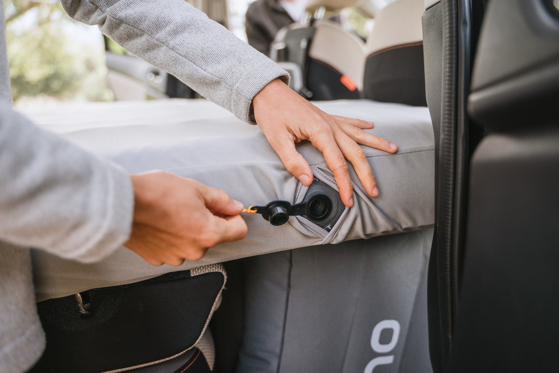 A person inflates an air mattress in the backseat of a car using a pump, with hands shown placing the pump nozzle into the mattress valve. The interior of the car and parts of a car seat are visible.