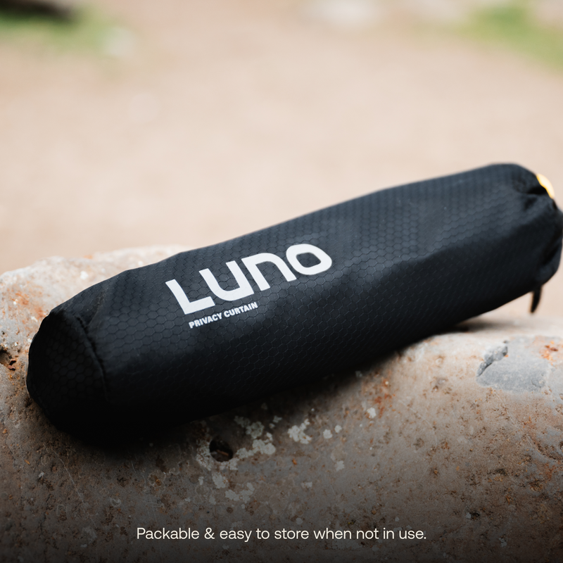 A black, cylindrical Car Privacy Curtain from Luno®, neatly packed and labeled with the name "Luno®," is placed on a rock. The text "Packable & easy to store when not in use." is displayed at the bottom of the image, promising sun-blocking protection for your ultimate sleeping space.