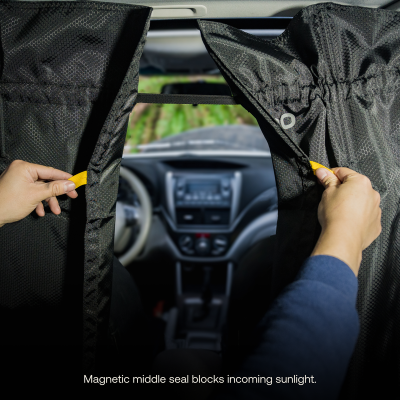 A person pulls the black Luno® Car Privacy Curtains closed inside a car to block incoming sunlight. The magnetic middle seal of these curtains ensures they stay shut, providing effective sun-blocking protection. The car's dashboard and windshield are visible in the background, creating the ultimate sleeping space.