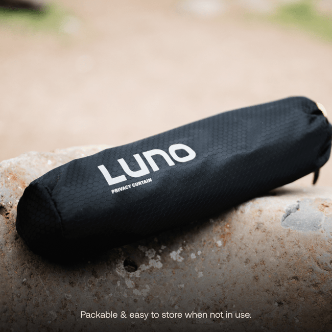 A black, cylindrical pre-owned Car Privacy Curtain bag from Luno HQ rests on a textured rock surface. The bag features a subtle hexagonal pattern and displays the Luno HQ logo along with "Privacy Curtain." In the background, a grassy area is slightly blurred, adding a natural touch to your travels.