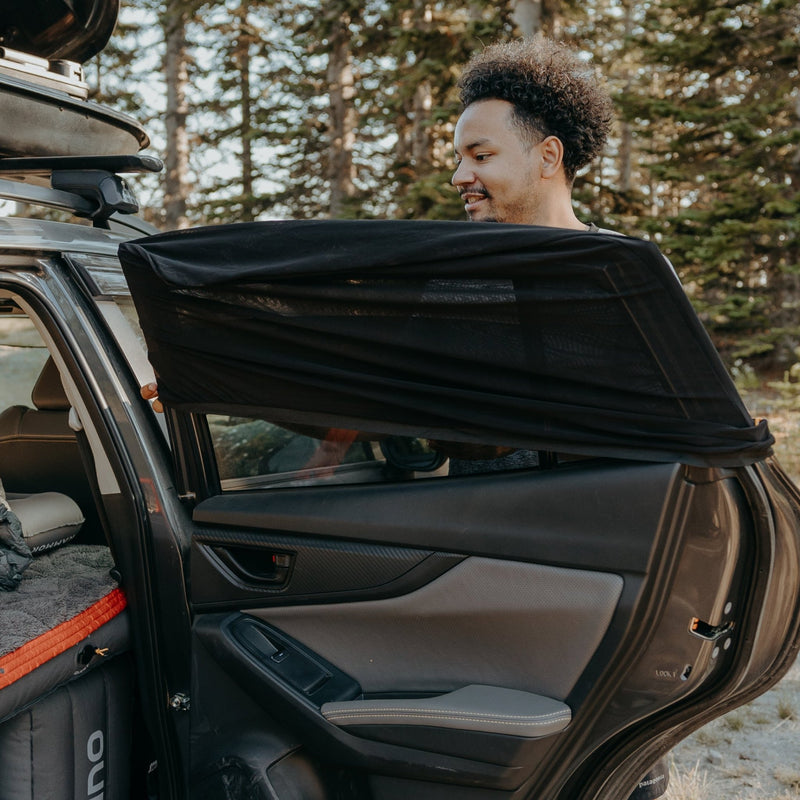 A man stands next to an open car door in a wooded area, installing a Luno HQ Car Window Screen over the rear window. Inside, some pre-owned gear is visible amidst the like-new condition of the tidy interior. Tall trees provide a serene backdrop.