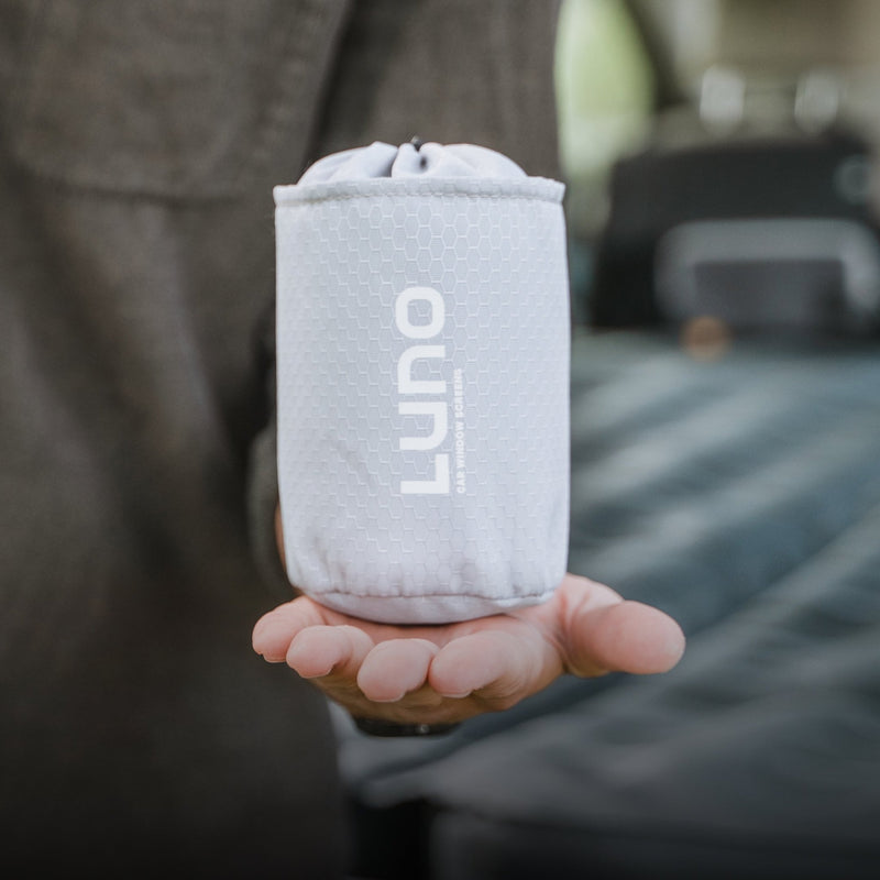 A person is holding a small, gray, cylindrical pouch with a hexagonal pattern and the "Luno HQ" branding printed in white. The light filters through the pre-owned Car Window Screens, revealing that the pouch looks like it's in pristine condition against the backdrop of a blurred interior.