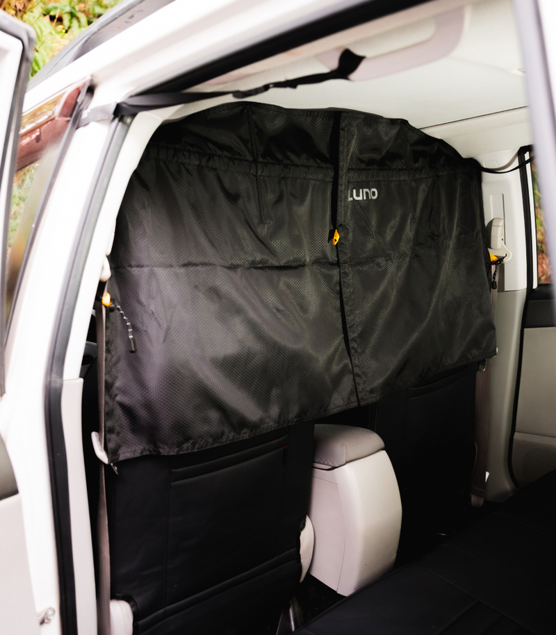 A black privacy screen labeled "Luno" is set up inside a white vehicle, covering the back seat area. The screen is attached to the interior roof and sides of the car and appears to divide the front seats from the back seats.