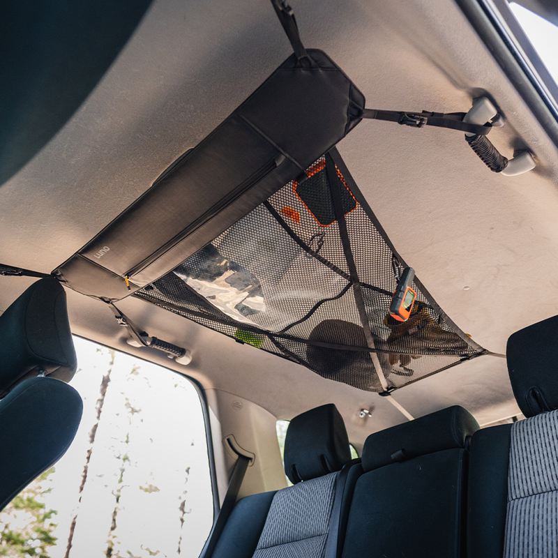 Interior view of an adventure mobile showcasing a Luno® Cargo Hammock attached to the ceiling above the back seats. Various items such as tools and small bags are stored on the portable gear shelf, maximizing storage space. The rear seats and part of the front seats are visible.