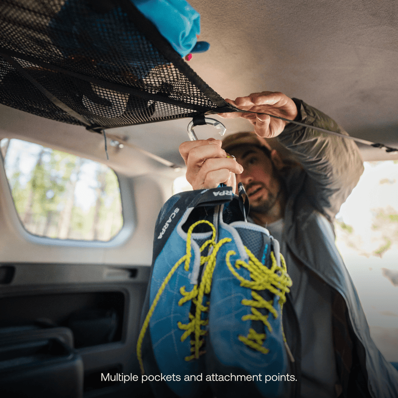 Using a carabiner, someone suspends a pair of Luno HQ blue climbing shoes with yellow laces from the car's overhead Cargo Hammock (Pre-Owned). The interior of the vehicle is visible, and the caption reads, "Smart storage: multiple pockets and attachment points.