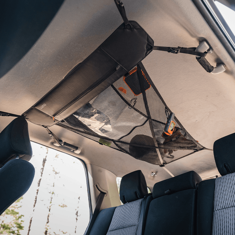 Inside the car, a Luno HQ Cargo Hammock (Pre-Owned) in black mesh is suspended from the ceiling, serving as a convenient portable gear shelf. This innovative storage option securely holds small items like a rope and a tool against the light background. Below, you can see the rear seats and headrests.