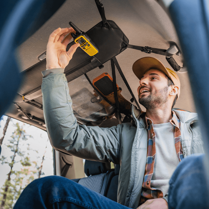 A man in outdoor attire reaches up to secure a yellow walkie-talkie in the Luno HQ Cargo Hammock (Pre-Owned) situated in the smart storage area of the vehicle's ceiling. He looks content, surrounded by forest scenery outside, as his neatly organized setup with portable gear shelves enhances his adventure.
