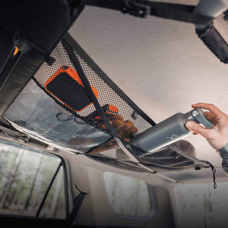 A person reaches for a reusable water bottle stored in a pre-owned Cargo Hammock by Luno HQ inside the vehicle. Nearby are an orange bag and other outdoor gear. Sunlight filters through the windows, revealing a forested scenery outside.