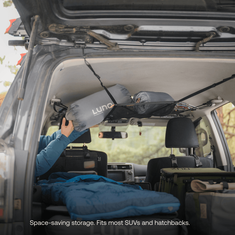 A person places a rolled-up Luno HQ sleeping bag into a pre-owned Cargo Hammock suspended from the SUV ceiling. The rear, packed with camping gear and a blue sleeping bag, showcases efficient storage. Text reads: "Space-saving storage. Fits most SUVs and hatchbacks.
