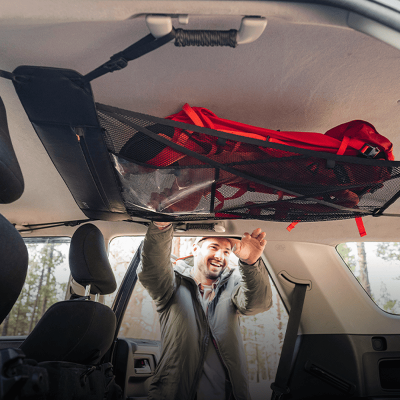 A person secures a red backpack in a Luno HQ Cargo Hammock (Pre-Owned) attached to the car's interior roof, with seats and windows in the background. The scene is outdoors, where trees are visible through the windows, showcasing this innovative portable gear shelf.