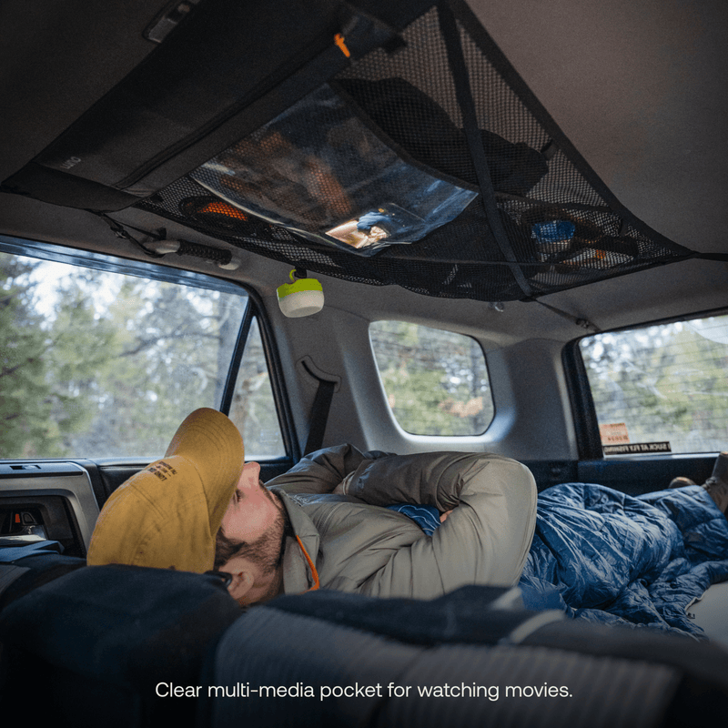 A person relaxes in the rear of a vehicle, gazing up at a multimedia device nestled in a Luno HQ Cargo Hammock (Pre-Owned) overhead. They're resting on a sleeping bag, donned in a yellow cap and jacket. The interior is softly illuminated, with trees visible through the windows.