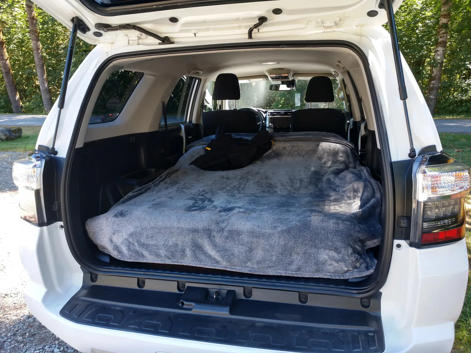 A white SUV with the rear hatch open, showcasing the back seats folded down and a large, neatly made bed with a grey blanket. The setup suggests the vehicle is used for car camping, offering a cozy sleeping space inside. Trees and a gravel path are visible in the background.