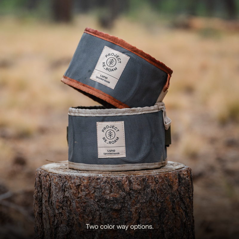 Two foldable, eco-friendly Collapsible Dog Bowls with "Luno HQ" labels, stacked on a tree stump in a forest setting. One bowl has a gray exterior with a brown rim, and the other has a beige rim. Text at the bottom reads, "Two color way options." Perfect for your portable pet needs!