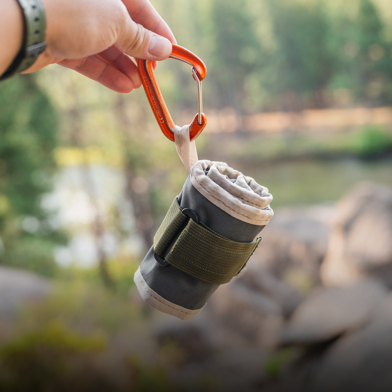 A hand is holding a rolled-up Luno HQ Collapsible Dog Bowl in green and gray, equipped with a carabiner clip, against a blurred outdoor backdrop of trees and a river.