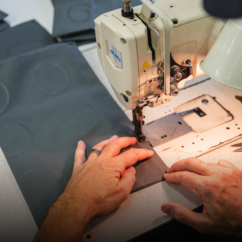 Close-up of a person using an industrial sewing machine to stitch fabric. The person's hands guide the material, which features circular patterns, under the machine's needle. The workspace is well-lit and includes a Luno HQ Collapsible Dog Bowl within easy reach.